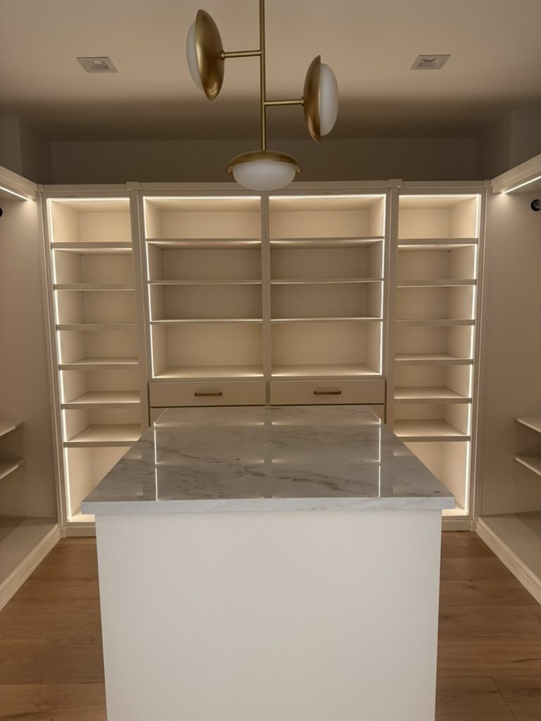 Master closet with LED tape lit along the shelving, a center island and a brass pendant light